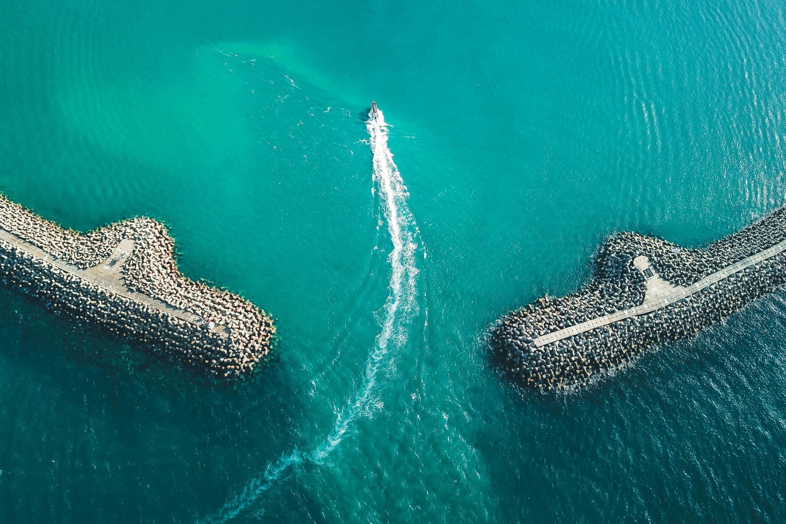 Drone shot capturing a boat traveling between two stone breakwaters in a serene ocean setting.